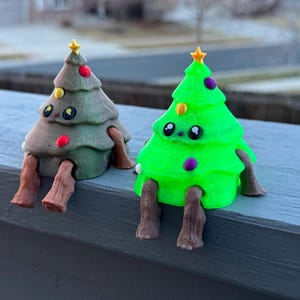 May include: Two whimsical Christmas tree figurines. One is green with purple and yellow ornaments, the other is brown with red and yellow ornaments. Both have smiling faces, star toppers, and brown legs, sitting on a gray surface.