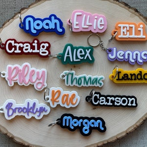May include: A collection of 13 colorful personalized keychains with names including Noah, Ellie, Eli, Craig, Alex, Jenna, Riley, Thomas, Landon, Rae, Carson, Brooklyn, and Morgan.