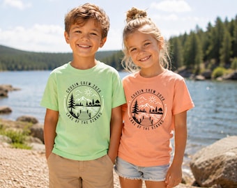 May include: A light green short-sleeve t-shirt and a peach short-sleeve t-shirt, each featuring a black circular graphic with a sun, mountains, trees, and a dock, with the text "COUSIN CREW 2026 LAKE OF THE OZARKS" printed in a circular border.