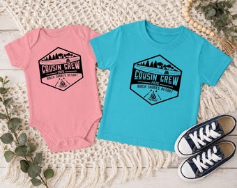 May include: A light pink baby bodysuit and a bright turquoise t-shirt, both featuring a black hexagonal graphic with a forest, lake, campfire, and the text COUSIN CREW 2026 BIRCH SHORES RESORT. Two navy blue and white canvas sneakers are placed nearby.