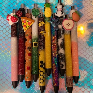 May include: A set of ten colorful pens with various decorative toppers, including a cow, a strawberry, a pizza slice, a shamrock, a bee, a butterfly, and a pineapple. The pens are all black and have a silver clip.