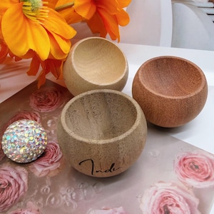 May include: Three small, round, bowl-shaped wooden objects with smooth, concave interiors. One bowl is light beige, one is medium brown, and one is dark brown with the word "Inde" printed in black script on the front exterior surface.