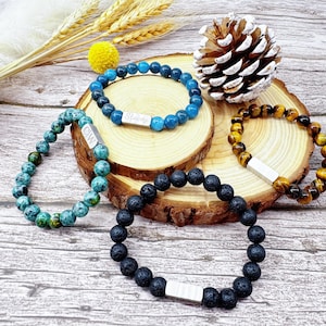May include: Four beaded bracelets in shades of blue, green, black, and brown. One bracelet has the name "Stacey" on a silver bar. The bracelets are displayed on a wooden slice with a pinecone and decorative elements.