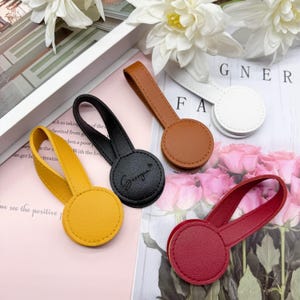 May include: A selection of leather curtain tiebacks in various colours: yellow, black, brown, white, and red. Each tieback features a loop handle and a circular base. The black tieback has the word "Georgia" printed on it.
