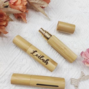 Pode incluir: Três atomizadores de perfume de bambu. Um atomizador está aberto, revelando um borrifador dourado. Um atomizador tem o nome "Leilah" gravado. Os atomizadores estão em uma superfície branca texturizada, com detalhes florais.