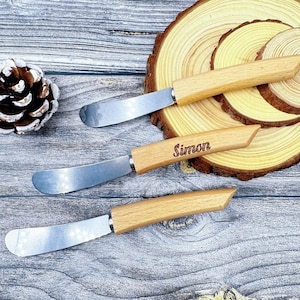 May include: Three butter knives with wooden handles and stainless steel blades. One knife has the name "Simon" engraved on the handle. The knives are displayed on a wooden surface.
