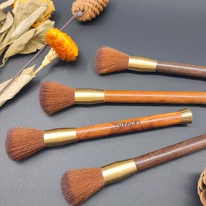 Peut inclure: Quatre pinceaux de maquillage avec des poils bruns, des manches en bois et des viroles en métal doré. Un pinceau porte le nom "Simon" gravé sur le manche. Les pinceaux sont disposés sur une surface sombre.