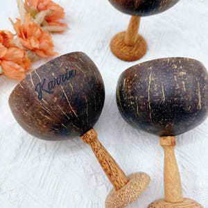 Peut inclure: Trois gobelets artisanaux en coquille de noix de coco avec des tiges et des bases en bois. Les coques brun foncé ont un aspect naturel et texturé, avec un gobelet affichant le nom "Karrin" en écriture noire. Les gobelets sont disposés sur une surface blanche.