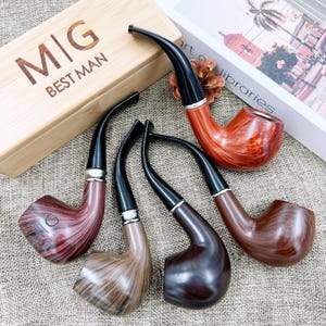 May include: Four wooden tobacco pipes with curved stems and black mouthpieces are displayed. The pipes vary in color, from light to dark brown, and are arranged near a wooden box with the text "M|G BEST MAN".