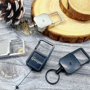 May include: A collection of keychains with carabiner clips. One keychain is black with the name "Giorgio" engraved. Another is silver with a rectangular design. The keychains are on a wooden surface.