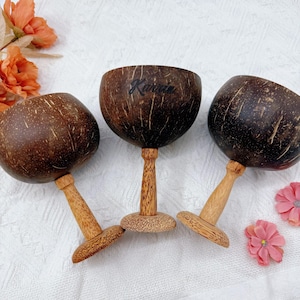 May include: Three handcrafted coconut shell goblets with wooden stems and bases. The dark brown shells are engraved with the name "Karrie" on one. The goblets are arranged on a white surface with pink flowers.