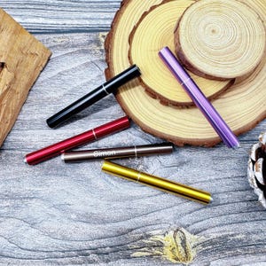 Puede incluir: Una variedad de bolígrafos metálicos delgados en negro, rojo, marrón, dorado y morado. Un bolígrafo está grabado con el nombre "Simon". Los bolígrafos están colocados sobre una superficie de madera con rodajas de madera.