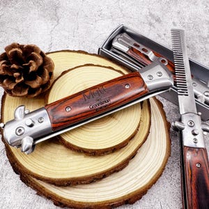 May include: A wooden-handled switchblade knife engraved with the name "Matt" and the word "Groomsman". The knife has a silver metal frame and is displayed on a wooden slice. A comb is also visible.
