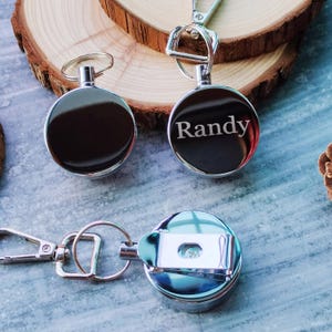 Può includere: Due portachiavi retrattili argentati con finitura nera e argentata. Un portachiavi ha il nome "Randy" inciso su di esso. L'altro portachiavi è vuoto.