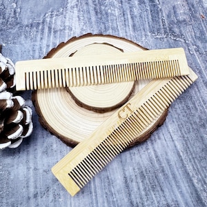 Könnte beinhalten: Zwei hellfarbene Bambuskämme mit feinen Zähnen werden auf einer Holzscheibe präsentiert. Ein Kamm ist auf dem anderen positioniert. Der untere Kamm hat die Initialen "CT" eingraviert. Der Hintergrund ist eine strukturierte, graue Oberfläche.