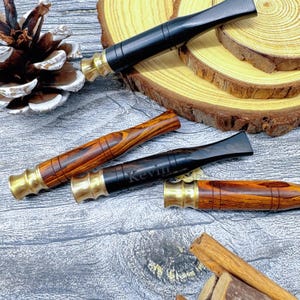 Può includere: Quattro bocchini per pipa in varie tonalità di legno, tra cui marrone scuro e rossiccio, con dettagli in ottone. Un bocchino è inciso con il nome "Kevin". Le pipe sono disposte su una superficie strutturata con fette di legno e una pigna.