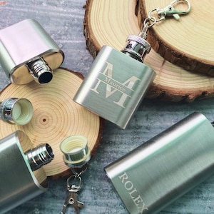 Op de afbeelding: Drie zilveren hipflasks met gegraveerde letters. Eén fles heeft de naam "Maurice" gegraveerd, een andere heeft het woord "Rolex" gegraveerd en de derde is simpel. De flessen staan op een houten oppervlak.