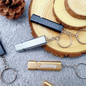Puede incluir: Primer plano de silbatos metálicos personalizados en plata, oro y negro, cada uno con un llavero. El silbato negro está grabado con "Anna", y el silbato dorado está grabado con "Jeremy". Los silbatos descansan sobre una superficie texturizada.