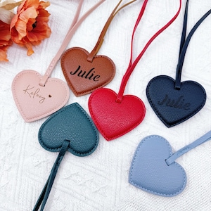 Può includere: Una collezione di etichette in pelle a forma di cuore in vari colori, tra cui rosa, marrone, rosso, blu navy, verde acqua e azzurro. Ogni etichetta ha un cinturino abbinato e presenta nomi personalizzati come "Julie" e "Kelsey" in corsivo. Le etichette sono disposte su una superficie bianca.
