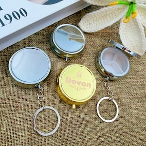 May include: A collection of round, metallic pill cases with keychains. One is gold with "Devon" printed on it, and others are silver. One case is open, revealing a white interior with three compartments.