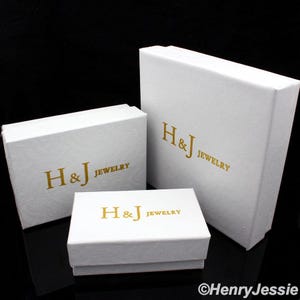 May include: Three white jewelry boxes of varying sizes. Each box has a textured surface and gold lettering that reads "H&J JEWELRY". The boxes are arranged on a black surface.