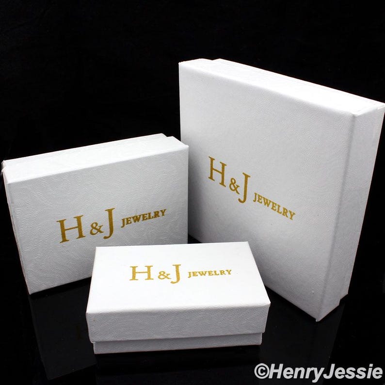 May include: Three white jewelry boxes of varying sizes. Each box has a textured surface and gold lettering that reads "H&J JEWELRY". The boxes are arranged on a black surface.