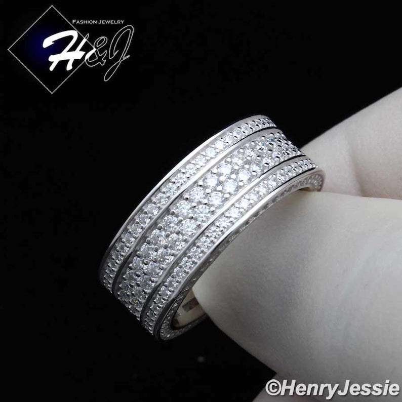 May include: A silver-toned ring with multiple rows of small, clear gemstones. The ring has a wide band design, with the gemstones set closely together. The ring is held against a white background. The ring is fashion jewelry.