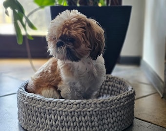 May include: A small, brown and white dog sits in a gray crocheted pet bed. The bed is round and has a white, fluffy lining.
