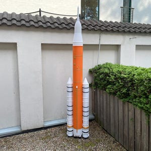 May include: A tall, orange and white model rocket stands outdoors. The rocket has a cylindrical body with white boosters and a pointed white nose cone. It is set against a backdrop of a white wall, a green hedge, and a wooden fence.