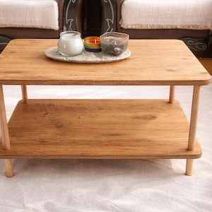 May include: A rectangular wooden coffee table with a lower shelf. The table is made of light-colored wood and has rounded corners. A decorative tray with candles and a glass container sits on top. The table is on a white rug.