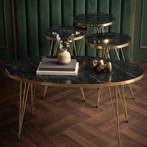 May include: A set of four nesting tables featuring black marble-patterned oval and circular tops with thin gold-toned metal legs. The largest oval table holds a small ceramic vase with dried stems, two stacked rectangular books, and a gold abstract sculpture.