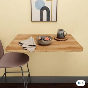 May include: A wall-mounted rectangular wooden table with a light brown grain finish. On the table sits a ceramic bowl filled with granola, blueberries, and strawberries, a folded beige linen napkin, and a two-toned grey mug on a wood slice coaster.
