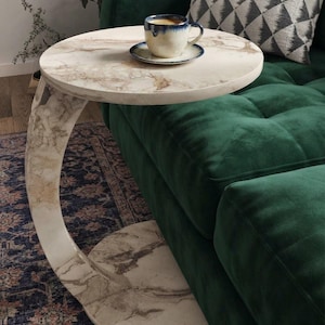 Può includere: Un tavolino a forma di C con una finitura a motivo marmorizzato bianco e beige. Il tavolo presenta un piano circolare e una base circolare abbinata collegati da una colonna di supporto curva. Una tazza da caffè in ceramica è sul piano.