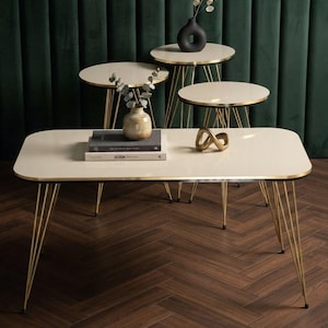 Peut inclure: Un ensemble de quatre tables avec des plateaux rectangulaires et circulaires blanc cassé et des pieds en métal doré. La table rectangulaire porte deux livres empilés et un petit vase avec de la verdure. Deux tables rondes portent un vase noir.
