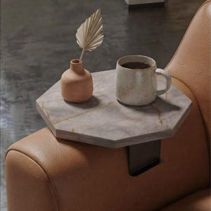 Puede incluir: Una mesa de reposabrazos octogonal con superficie de mármol gris claro y abrazadera de metal oscuro. Sobre la mesa hay un pequeño jarrón color melocotón mate con una hoja de palma seca y una taza de cerámica moteada de color claro.