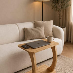 Peut inclure: Une table d'appoint en bambou clair en forme de C avec un plateau et une base rectangulaires, reliés par un support incurvé. Un ordinateur portable gris foncé, un smartphone noir et une tasse en céramique beige sont posés sur la table.