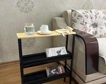 Op de afbeelding: Een zwarte metalen bijzettafel met een houten blad en twee planken. De planken zijn bekleed met zwarte stof. Er staan twee boeken op de planken, een met de titel "Kafka" en de andere met de titel "Sehille". Op de bovenste plank staan een glas water en een kopje koffie.