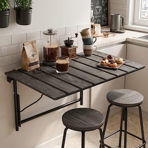 Puede incluir: Una mesa plegable de pared de madera oscura con dos taburetes a juego, que incluye una prensa francesa, un vaso de café, un pequeño molinillo de café de madera, una pila de tazas de cerámica y una bandeja de pasteles sobre una pared de azulejos.
