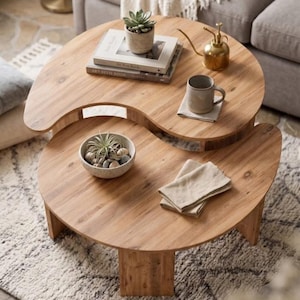 Può includere: Un set di due tavolini da caffè a incastro realizzati in legno chiaro con una forma curva ispirata allo yin e yang. Il tavolo superiore ospita una piccola pianta grassa in vaso, due libri impilati, un nebulizzatore in metallo dorato e una tazza in ceramica. Il tavolo inferiore contiene una ciotola di pietre con una piccola pianta e tovaglioli di stoffa piegati.