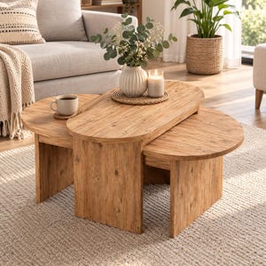 May include: A set of two oval-shaped wooden nesting coffee tables with a natural wood grain finish. The tables feature flat tops and solid panel legs. A white ribbed vase with greenery and a white candle sit on the top table surface.