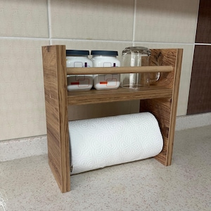 Könnte beinhalten: Ein Holz-Papierhandtuchhalter für die Arbeitsplatte mit zwei Ablagen. Die obere Ablage bietet Platz für zwei weiße Gläser und ein klares Glas. Die untere Ablage bietet Platz für eine Rolle weißer Papierhandtücher.