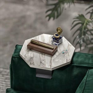 May include: An octagonal white marble sofa arm tray with a dark metal clamp. On the surface sits a brown wooden pen holder with a metallic pen, a square glass inkwell with a brass lid, and a silver-toned metal bookmark with a turquoise stone.