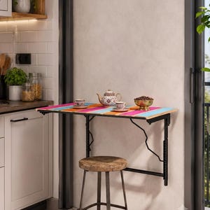 May include: A wall-mounted folding table with a multicolored striped wooden top and black metal support brackets. A round wooden stool with metal legs sits underneath. On the table are a patterned teapot, two matching cups, and a bowl of coffee beans.