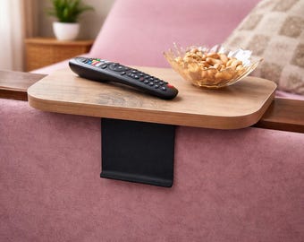 Peut inclure: Un plateau d'accoudoir rectangulaire en bois avec une finition grain de bois brun clair et une base de serrage en métal noir. Le plateau supporte une télécommande noire et un bol en verre transparent rempli de noix, posé sur un accoudoir rose.