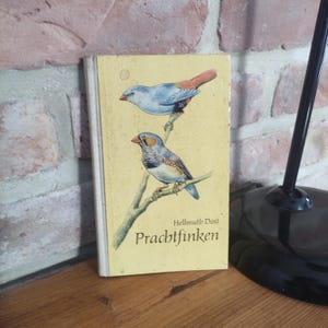 Könnte beinhalten: Ein Vintage-Buch mit dem Titel "Prachtfinken" von Hellmuth Dost, das Illustrationen von zwei bunten Vögeln auf einem Ast vor gelbem Hintergrund zeigt. Das Buch lehnt an einer Backsteinmauer.