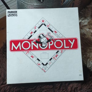 Könnte beinhalten: Monopoly-Brettspielbox. Das rot-weiße Logo zeigt eine Figur mit Zylinder und Gehstock. Die Box ist weiß mit einer rautenförmigen Spielbrettkontur.