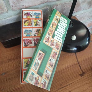 Könnte beinhalten: Vintage Domino-Set mit einer grünen Schachtel und Illustrationen von Zeichentrickfiguren auf den Dominosteinen. Auf der Schachtel steht "DOMINO" in weißen Buchstaben. Die Dominosteine befinden sich in einer weiß-orangenen Schachtel.