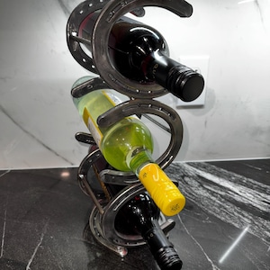 May include: A wine rack made of three stacked horseshoes, holding three bottles of wine. The rack is made of metal and has a rustic look. The bottles are a red wine, a white wine, and a red wine.