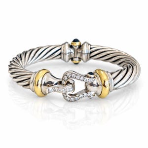 Puede incluir: Una pulsera de estilo cable hecha de plata de ley y oro amarillo, con un cierre en forma de herradura adornado con pequeños diamantes pavé transparentes y una piedra preciosa azul oscuro en la bisagra.