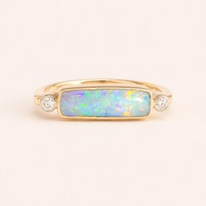May include: A gold ring with a rectangular opal displaying iridescent blues, greens, and yellows. Two small, round diamonds are set either side of the opal. The ring has a polished finish.